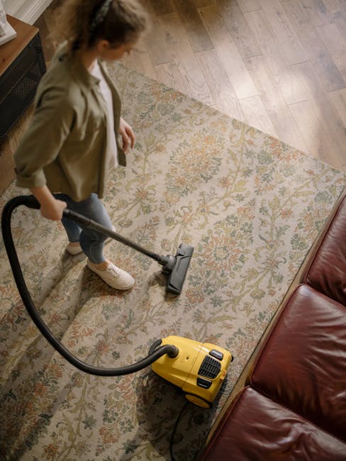 A woman vacuuming an ornate, patterned area rug with a yellow canister vacuum cleaner in a living room. The rug features intricate floral motifs in shades of beige, green, and orange, positioned next